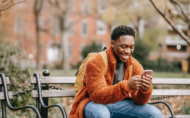 Man kijkt op zijn smartphone terwijl hij op een bankje in een park zit