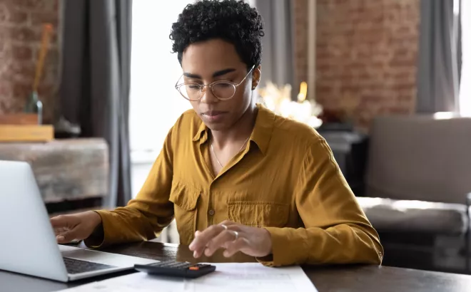 Vrouw doet een berekening op een rekenmachine terwijl ze op haar laptop werkt